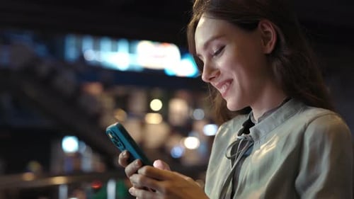 Beautiful Caucasian Smiling Girl Walks Down the Night Street and Texts Smiling at Her Phone