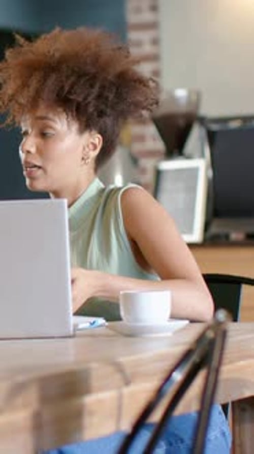 Vertical video: Reacting to message, woman in sleeveless top and jeans typing at cafe table, laptop