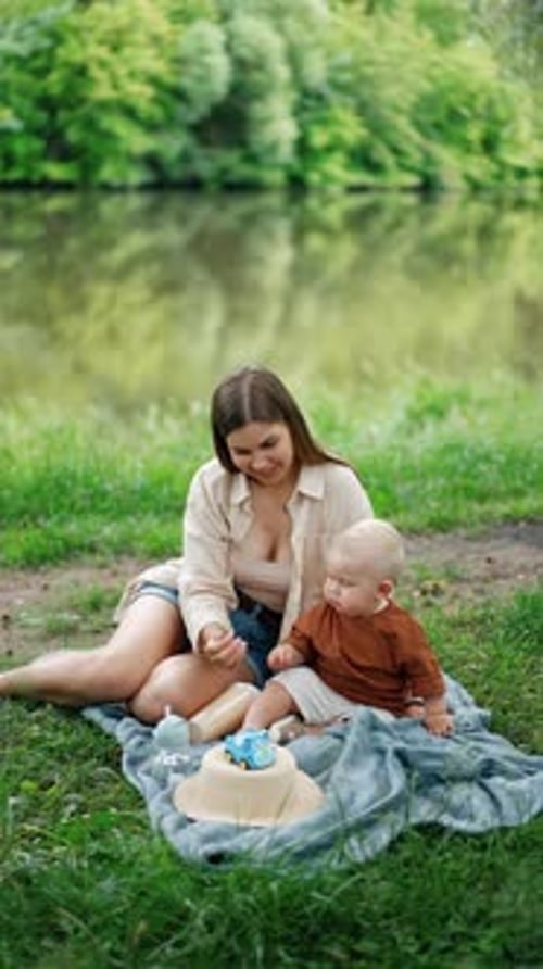 Mother and child sitting on the plaid near the river resting. Mom gives a leaf to her baby boy.
