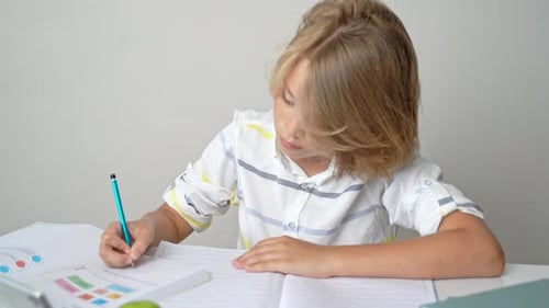 Middle School Smiling Student Boy Sitting at Desk Studying Writing Book Homework and Tablet at Class