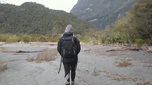 Girl in hiking gear walking along beach surrounded by lush green forrest and mountains