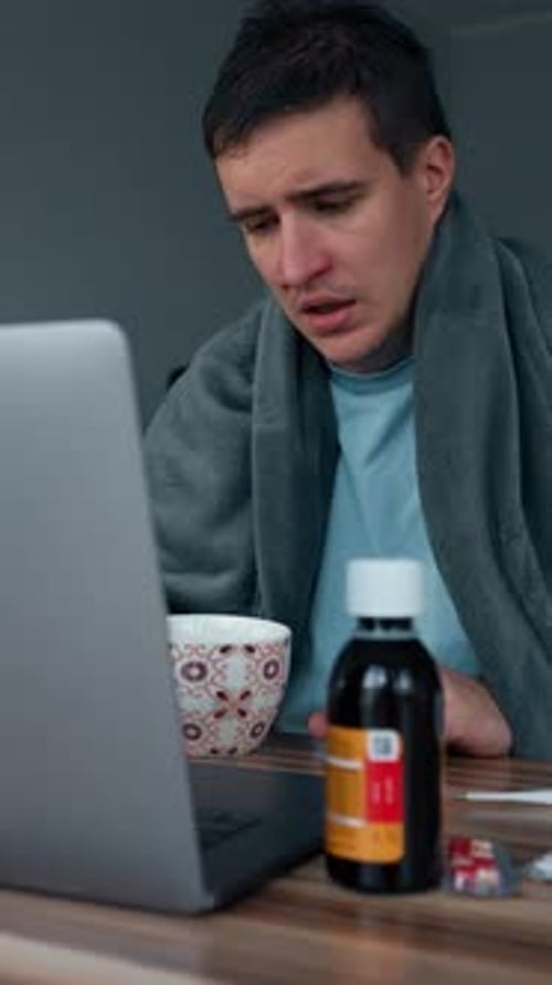 Vertical of Sick Man Drinks Tea Watching Online Courses on Laptop