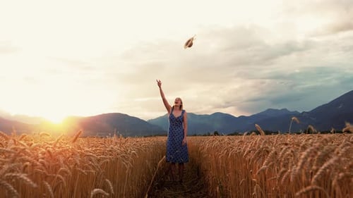A young woman stands in the middle of the wheat field and throws her hat into the air.
