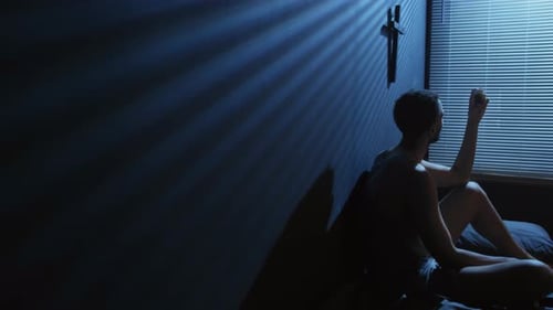 Young Adult Sits on Bed in Dark Room