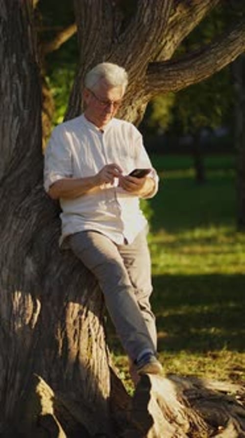 Man Using Mobile Phone Leaning Against Tree in Park