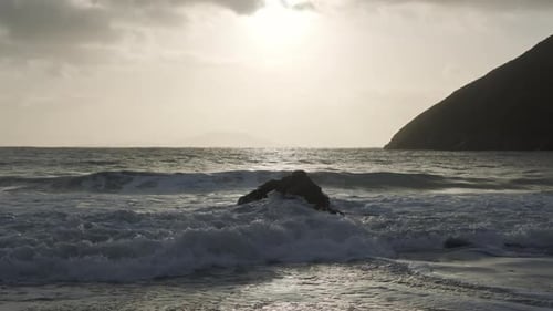 One strong boulder lying on a remote beach, waves crashing into it causing foamy waves and spray in
