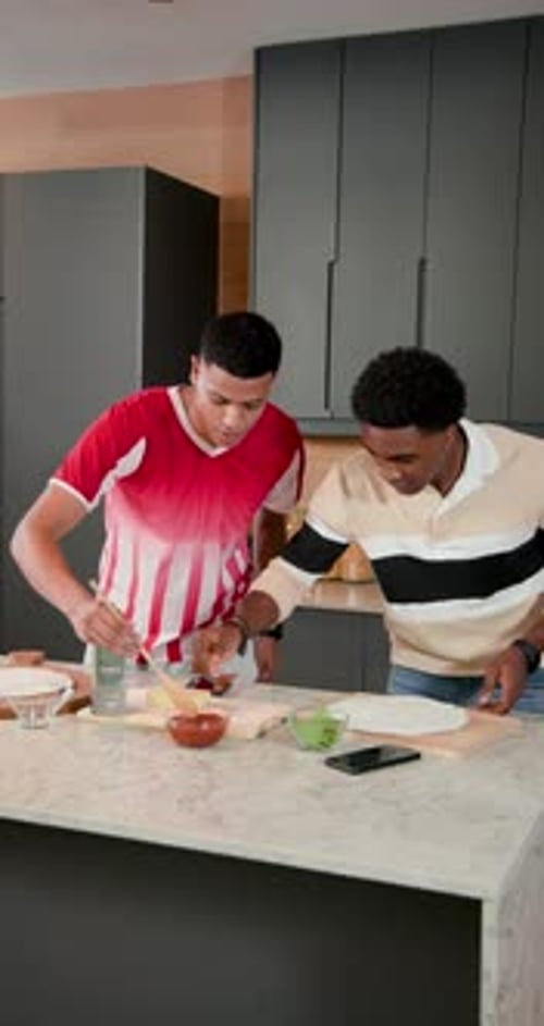 Vidéo verticale : amis de sexe masculin lisant une recette au téléphone étalant de la sauce, du fromage sur de la pâte sur le bar de la cuisine