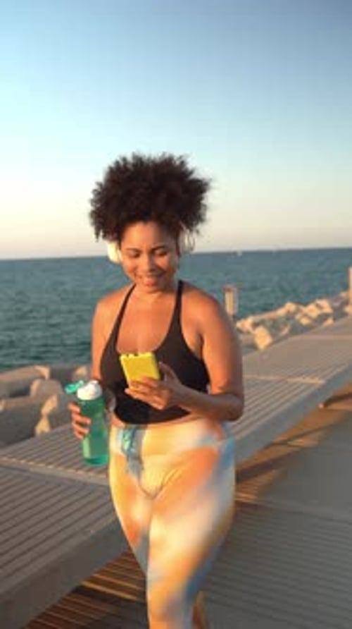 Plus-size Hispanic woman listening music and using smartphone while training outdoors on pier