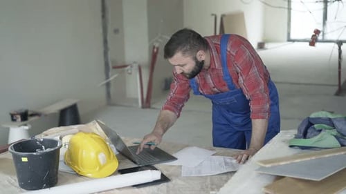 Bearded Man Reviews Construction Plans on Laptop Indoors