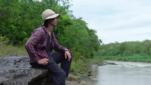 Ein Mann sitzt auf einem Felsen neben einem malerischen Fluss, umgeben von üppigem Wald