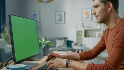 Handsome Man Sitting at His Desk at Home Uses Personal Computer with Mock-up Chroma Key Screen.