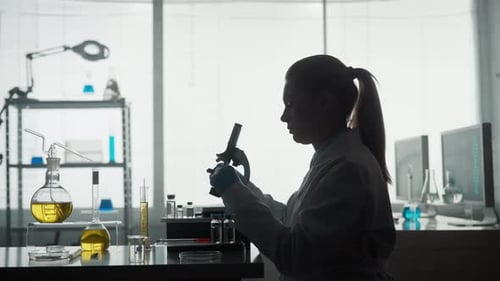 Side View of a Dark Silhouette of a Female Scientist Looking Under a Microscope Doing an Analysis of