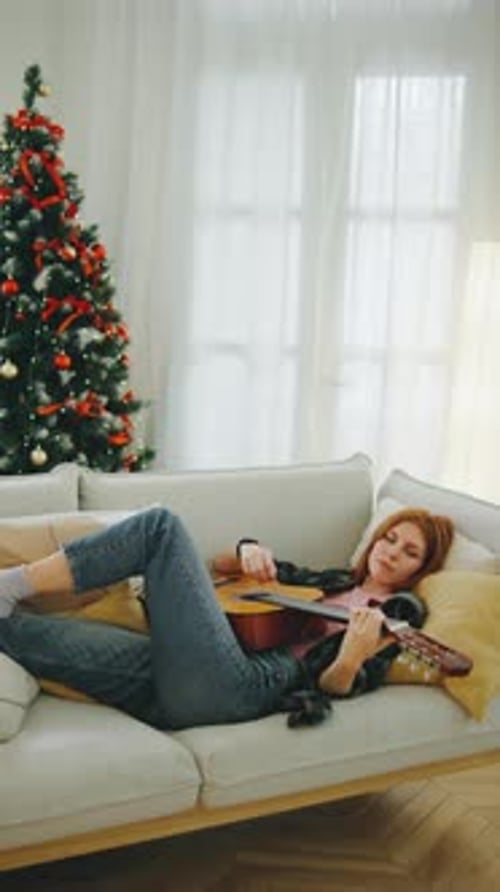 Young Woman Relaxes on a Sofa Playing Guitar Beside a Beautifully Decorated Christmas Tree During