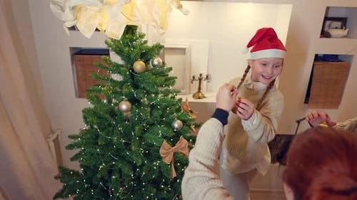 Cute Caucasian Girl is Decorating Christmas Fir Together with Her Mum and Hanging Ball on It