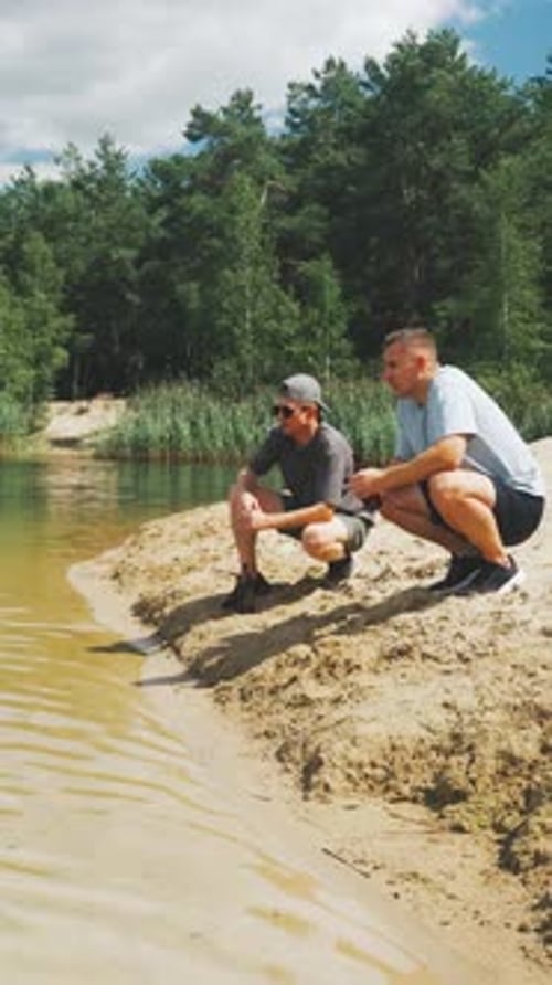 Relaxed Friends Sitting By Tranquil Water Two Companions Lounge Calmly on Sandy Riverbank Under Blue
