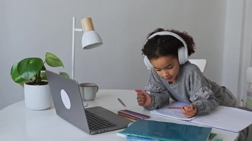 Primary School African American Student Child with Headphones Hand Writing in Book Using Laptop