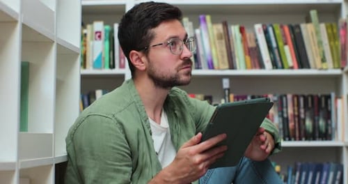 Young Man in Glasess Sitting on the Floor Near Shelves with Book in the Library Uses Digital Tablet