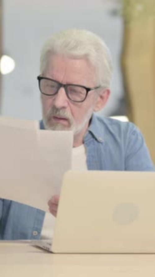 Senior Man Looks at Documents with Laptop
