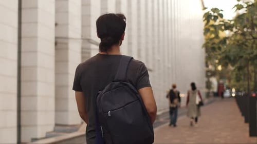 View From Behind of a Young Man with a Backpack Walks Alongside the Building with a Smiling Happy