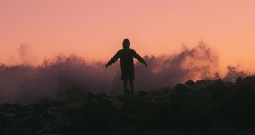 Teenager throws hands up as wave crashes against jetty causing a large spray of water in the backgro