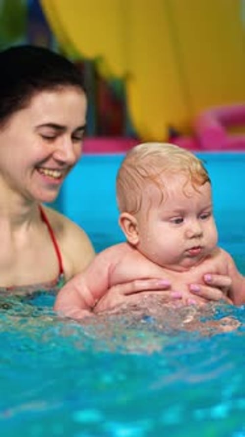 Adorable chubby baby is in the swimming-pool. Brunette woman teaching the infant to swim.