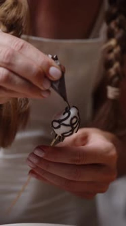 Vertical. The woman is decorating the strawberries in white chocolate with intricate patterns
