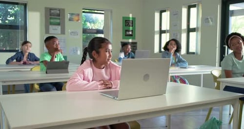 Female child in pink top turning to teacher entering from right in classroom, listening at laptop