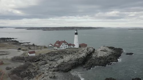 Drone shot, of a lighthouse, on a rocky coastline, during a cloudy day, flying to the right.