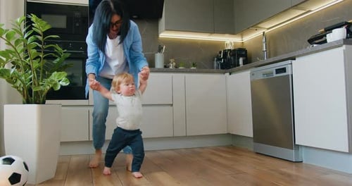 Caucasian cute little baby boy child learn to walk with mom in house. Mother holding hands baby