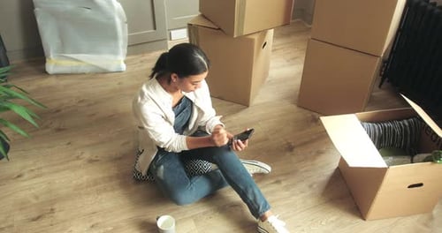 Woman Using Smartphone Surrounded by Moving Boxes