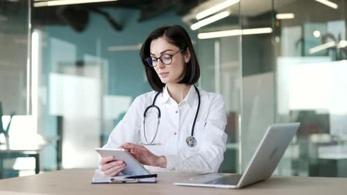 Female doctor in white coat using digital tablet sitting at workplace in office in modern hospital
