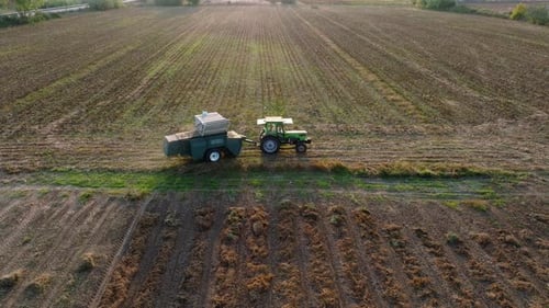 Aerial: Side follow of a tractor with trailer in an empty field