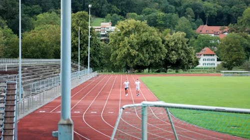 Two runners do their training laps on running track in front of empty stands on campus, wide shot