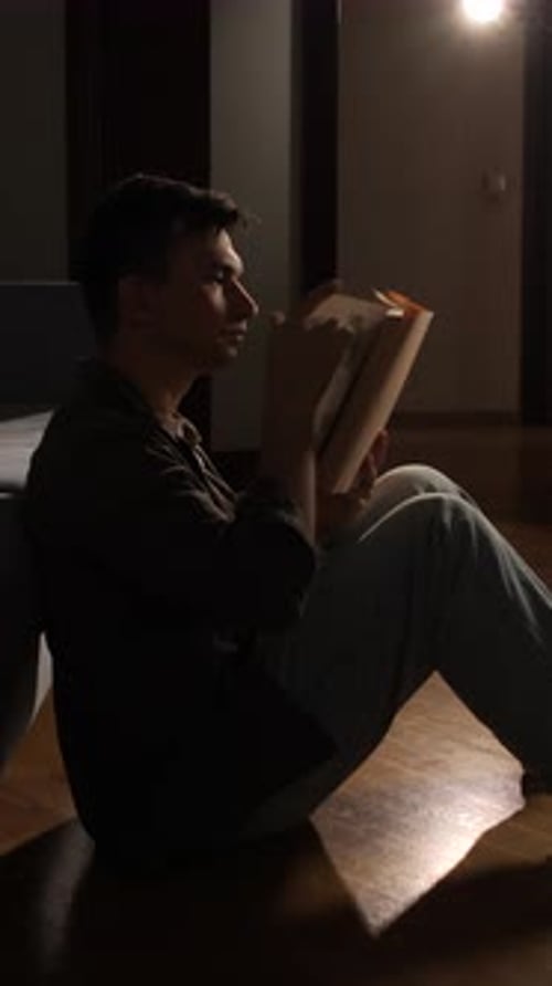 Vertical Video Young Man Sitting on a Wooden Floor Deeply Engrossed in Reading a Book Under a Soft