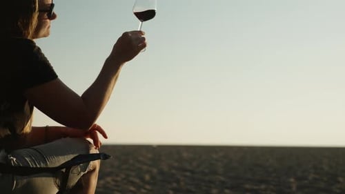 A romantic date on the beach by the sea, a young woman looks at the sunset through a glass of wine.