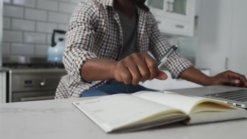 Midsection of african american man working at home in kitchen writing and using laptop