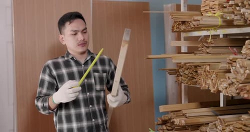 Man Measures Wood Plank with Tape Measure in Workshop
