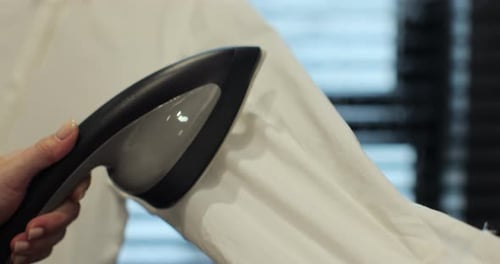 Unerkennbare Hand, die den Dampfgarer auf dem weißen Damenhemd benutzt, um Falten zu Hause zu glätten