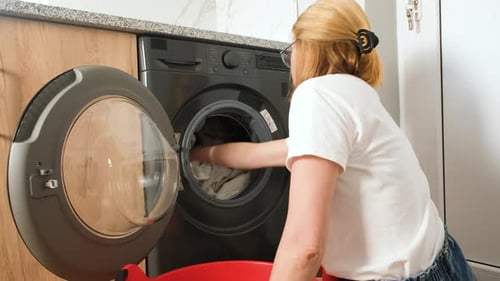 Young Woman Loading Laundry Into a Frontloading Washing Machine at Home She Kneels By a Red Basket