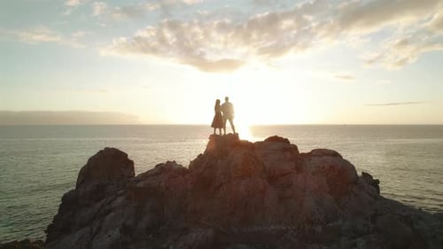 Lovers stand on a top of rocky cliffside by the ocean, woman and man embracing and overlooking sunse