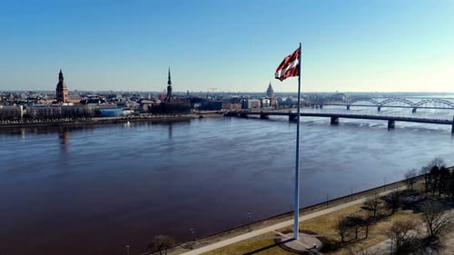 Ein Fahnenmast steht vor einer Stadt und einem Gewässer, dem Fluss Daugava. Die lettische Flagge ist rot
und