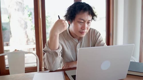 Focused serious Asian watching online learning course on laptop and taking a note