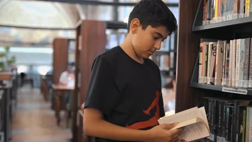 Boy Reads Book Next To Bookshelf In Library