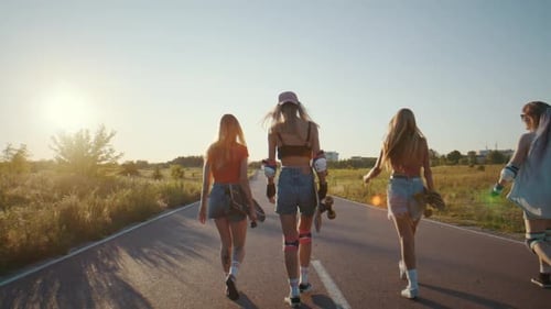 Women Stroll Along Sunsetlit Paved Road with Surfskates in Hands