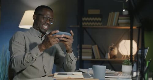 Hombre afroamericano despreocupado jugando a un videojuego en línea con un teléfono inteligente disfrutando de la Aplicación móvil en la oscuridad
