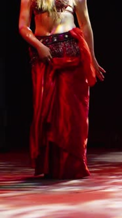 Vertical video. A woman performs a belly dance on stage. Red costume and lighting, spectators