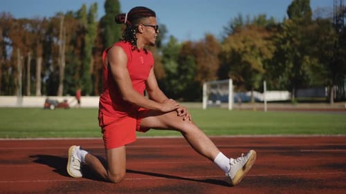 A Male Athlete Stretches His Knee Joint While Standing Outdoors on a Sunny Autumn Day on a Stadium