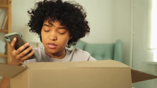 Young Woman with Phone Opens Delivery Box at Home