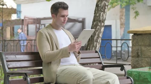Young Man using Tablet while Sitting Outdoor on a Bench