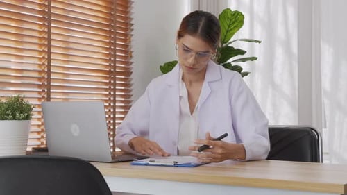 Young woman doctor working while looking document on clipboard and using laptop computer at clinic.